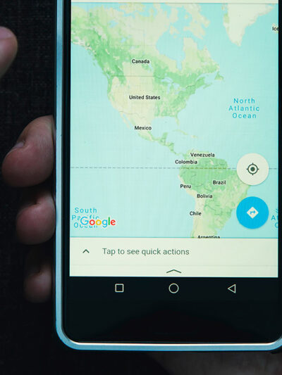 One hand holding a cell phone while the other hovers with index finger extended over a smart phone showing a map of North and South America.