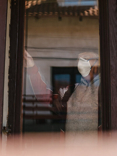 An elderly person wearing a mask looks out a brown wood frame window.