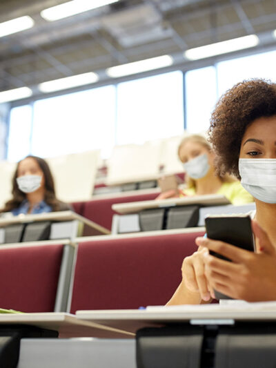 A girl sits in a college lecture hall, wearing a mask and scrolling through phone in hand.