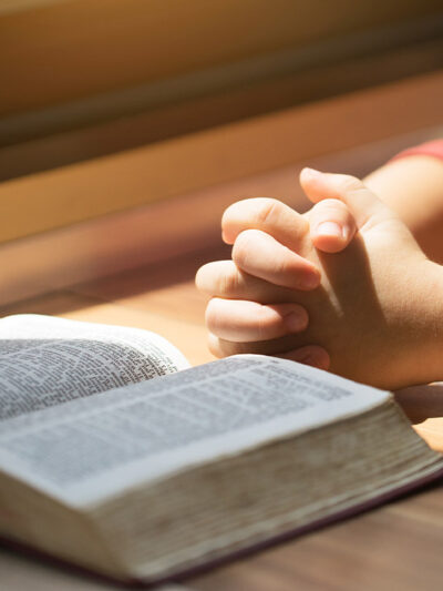 A boy whose hands are folded in prayer in front of a Bible on a desk.