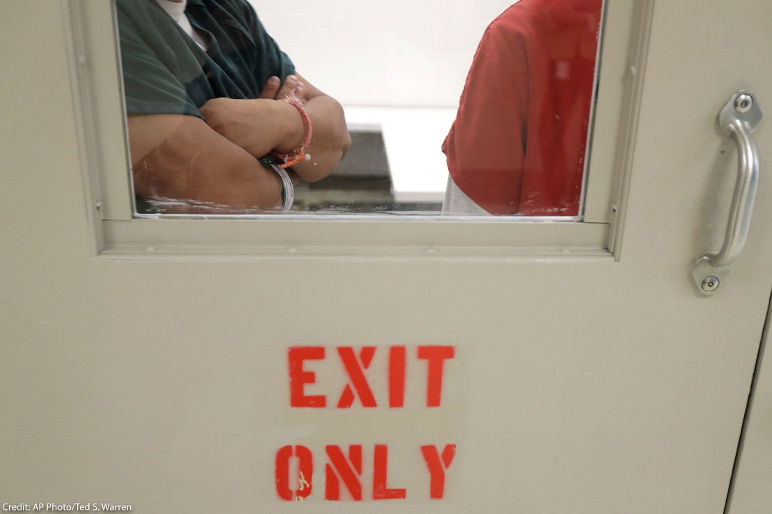 Two detainees seen waiting through a window behind closed doors.