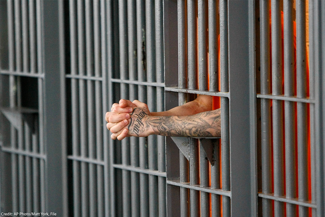 A prisoner holding his extended and clasped hands between the bars of his cell.