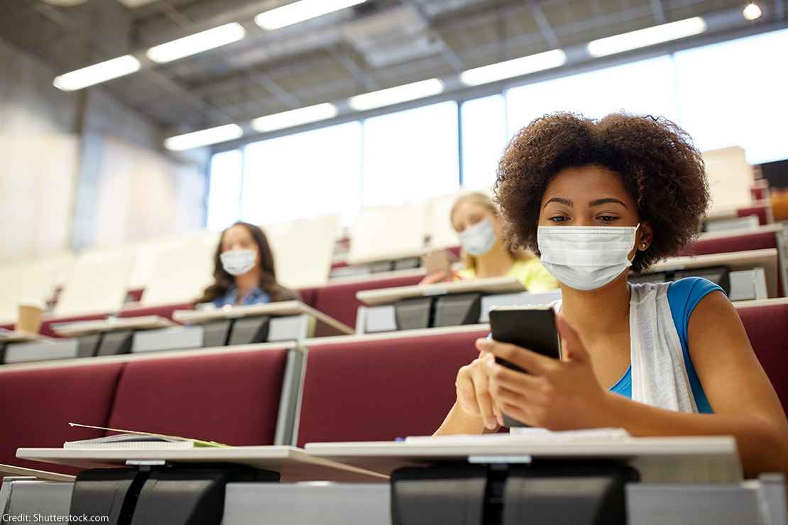 A girl sits in a college lecture hall, wearing a mask and scrolling through phone in hand.