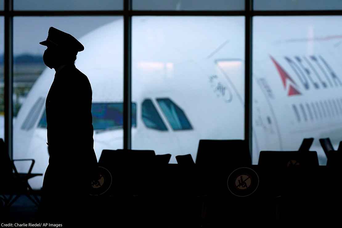 The silhouette of a pilot wearing a facemask walking past a window with a Delta plane outside.