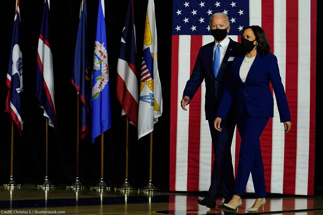 President-Elect Joe Biden and Vice President-Elect Kamala Harris arrive on stage at campaign event with American flag in background.