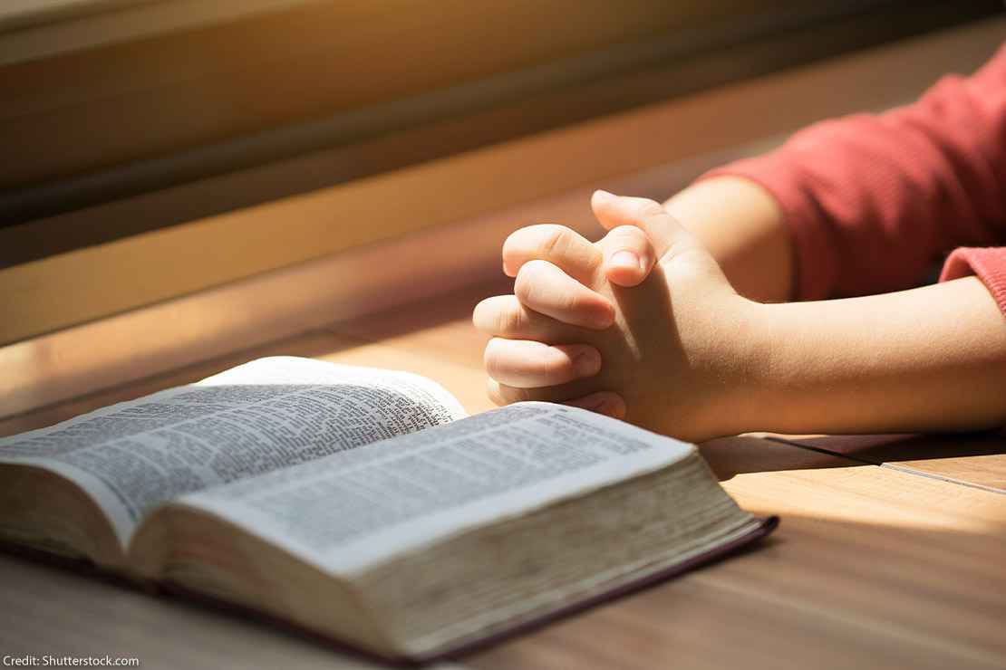 A boy whose hands are folded in prayer in front of a Bible on a desk.