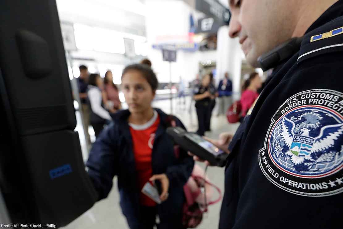 A passenger using a facial recognition kiosk in the background with a U.S. Customs and Border Protection officer watching in the foreground.