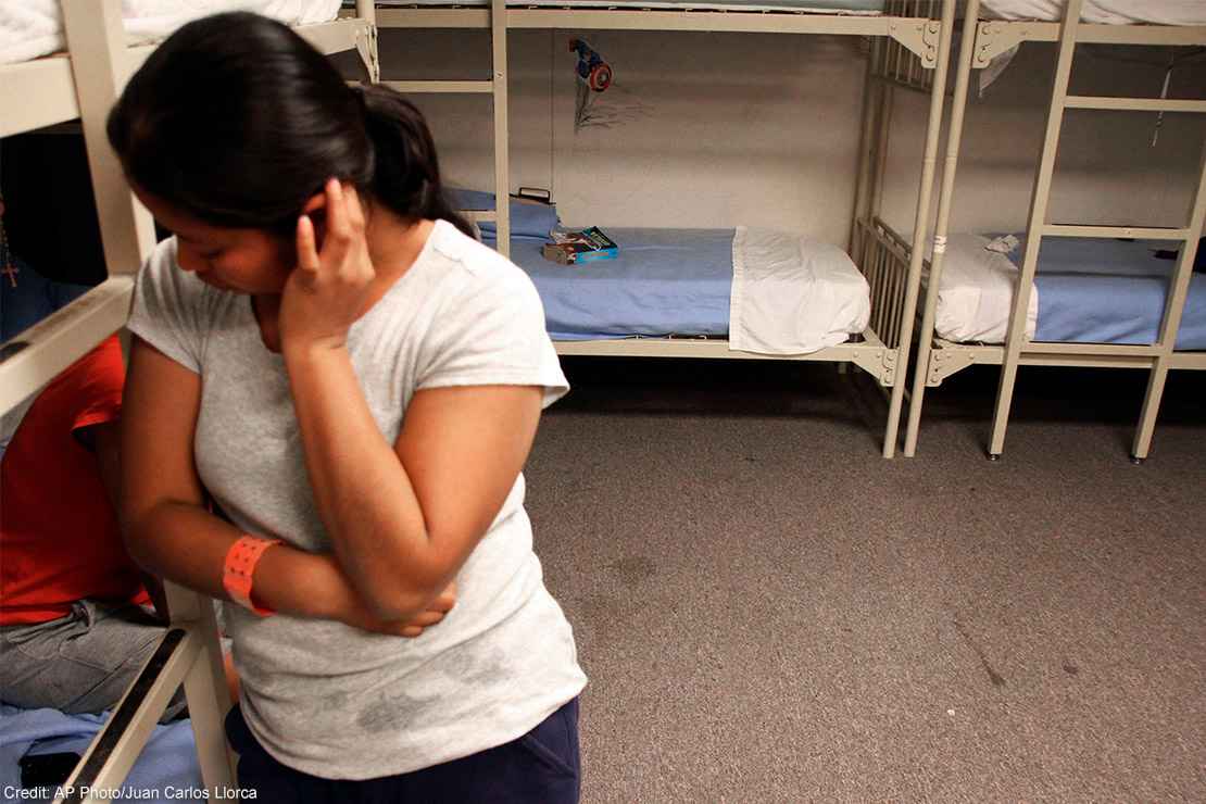 A migrant detainee stands facing away from bunks.