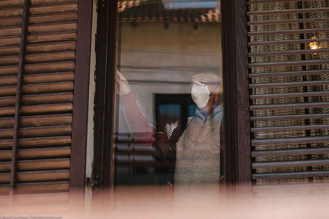 An elderly person wearing a mask looks out a brown wood frame window.