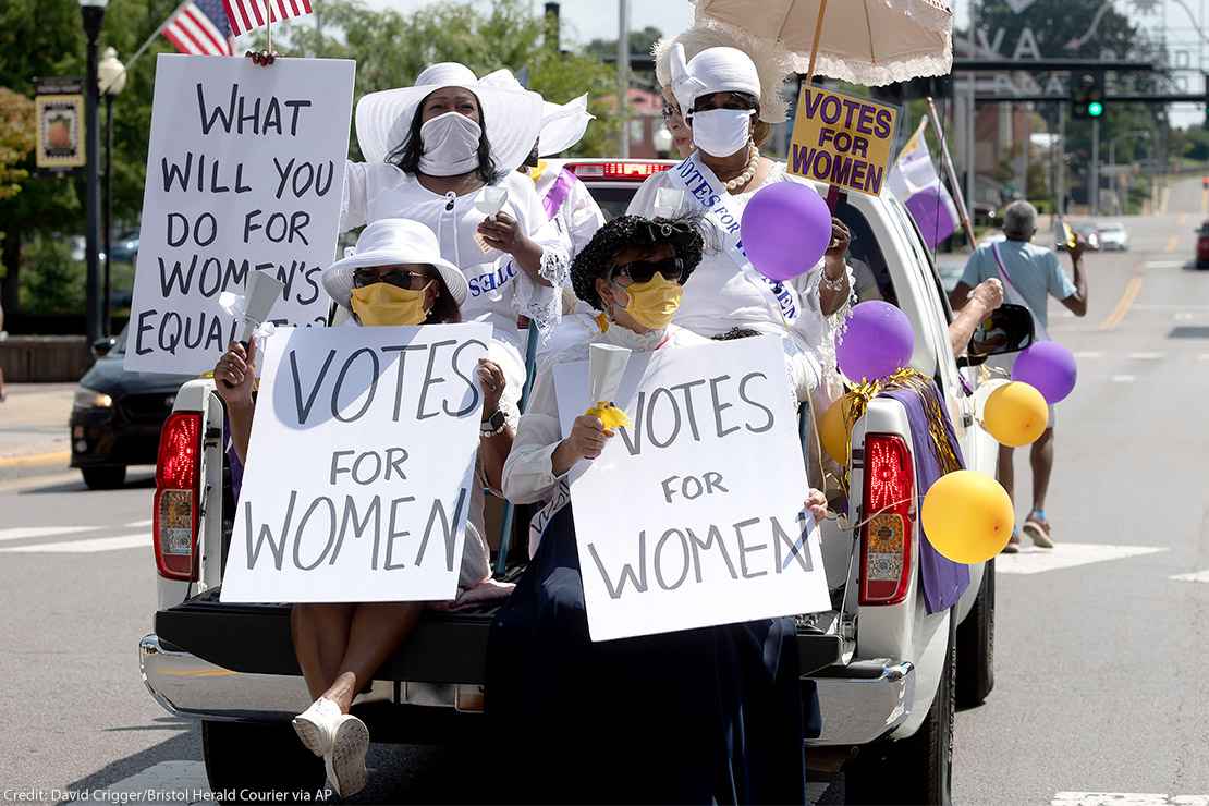 Demonstrators in period clothing with signs advocating for women's suffrage.