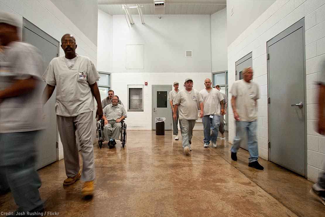 Prisoners are seen walking in a hallway in a prison, one in a wheelchair