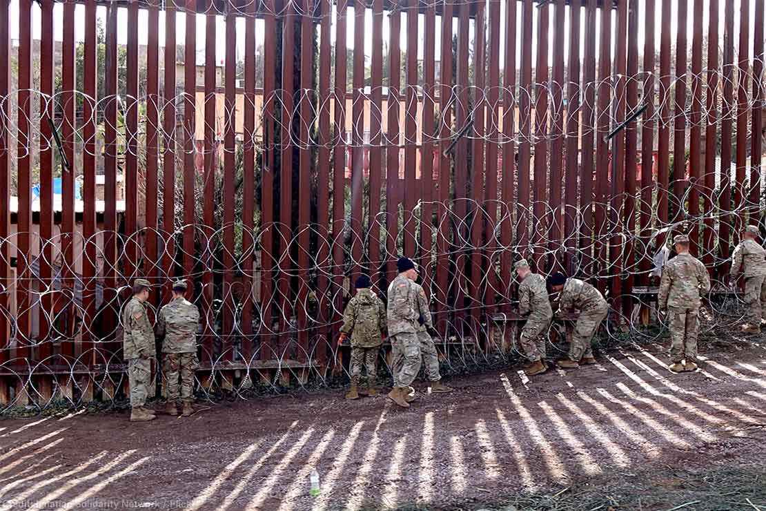 US military members are seen next to a red metal and barbed wire fence at the US border